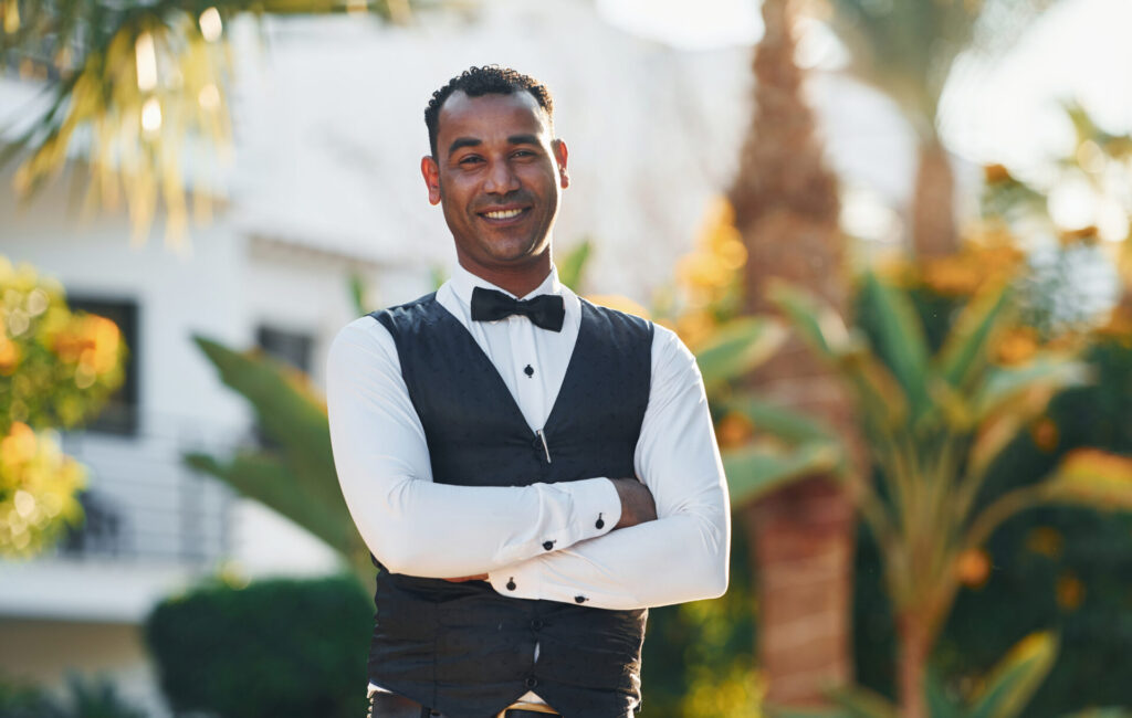 Smiling man in a white shirt with black vest and bow tie standing outdoors with crossed arms.