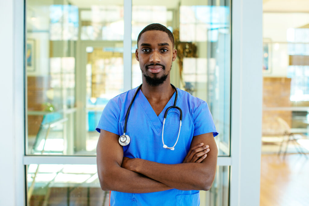 Confident young male healthcare worker in blue scrubs with stethoscope, standing with arms crossed indoors.