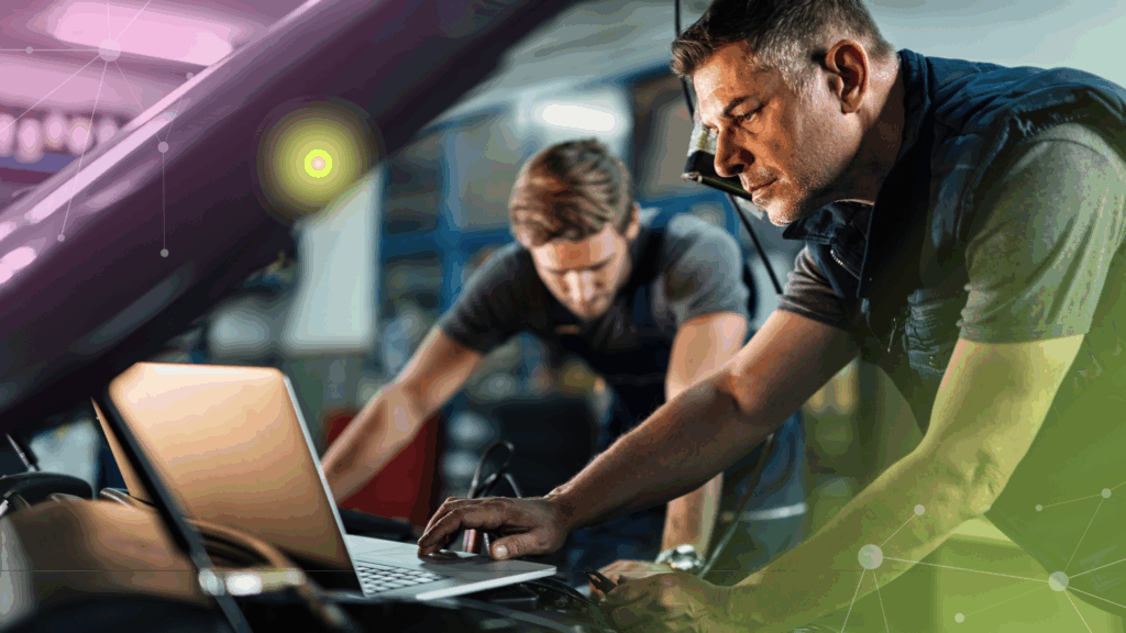 Two men working on a car engine using a laptop in a garage or workshop.