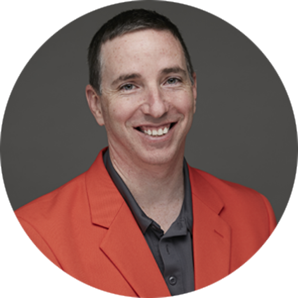 Smiling man with short dark hair wearing an orange blazer and dark shirt against gray background.