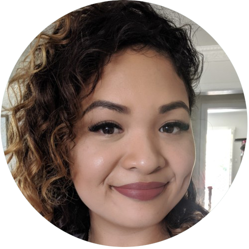 Woman with curly hair smiling, wearing makeup, with a softly lit room in the background.