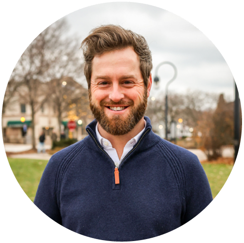 Smiling bearded man with curled hair wearing a navy pullover standing outdoors in a park.