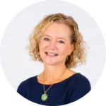 Smiling woman with curly blonde hair wearing a blue top and green pendant necklace.