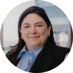 Smiling woman with dark hair wearing a blue collared shirt and black blazer in an office setting.