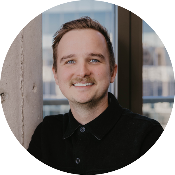 Smiling man with mustache wearing a black shirt standing indoors by a concrete column and window.