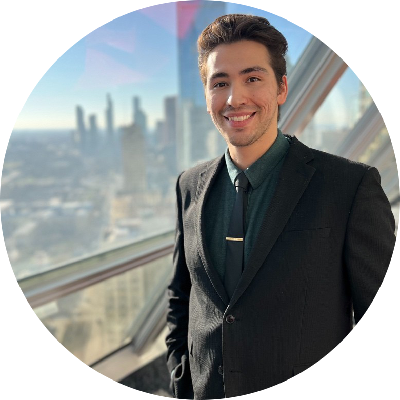 Smiling man in black suit and tie standing by window with city skyline in background.