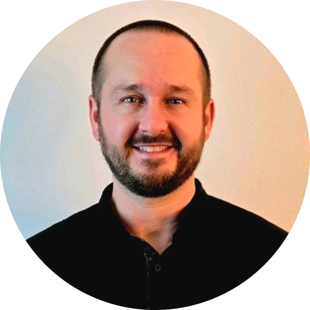Smiling man with short hair and beard wearing a black shirt against a plain light background.