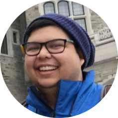 Smiling person wearing glasses, a blue knit hat, and a blue jacket in front of a stone building.