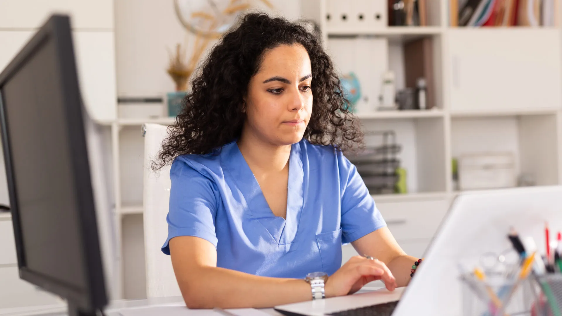 Nurse in blue scrubs focused on typing on a laptop in a bright office with shelves in the background.