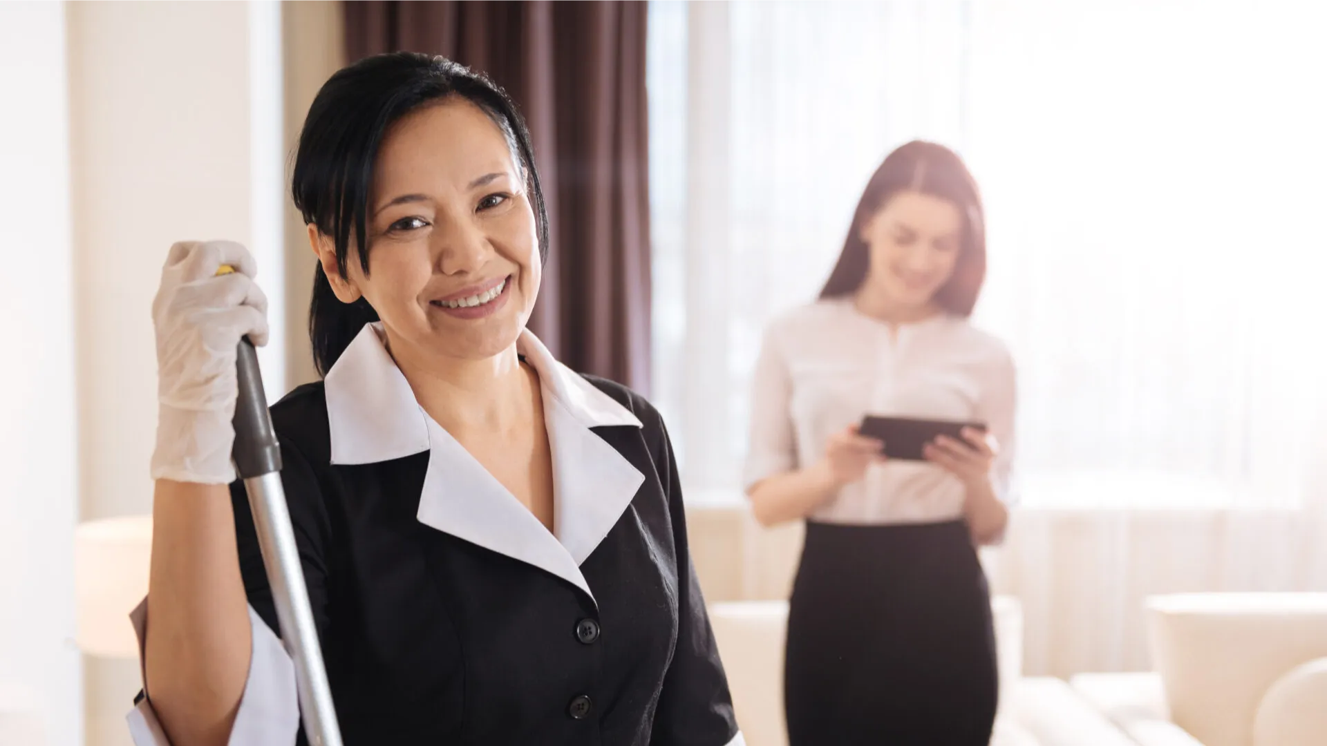 Smiling hotel maid holding a mop in front of a woman using a tablet in a room.