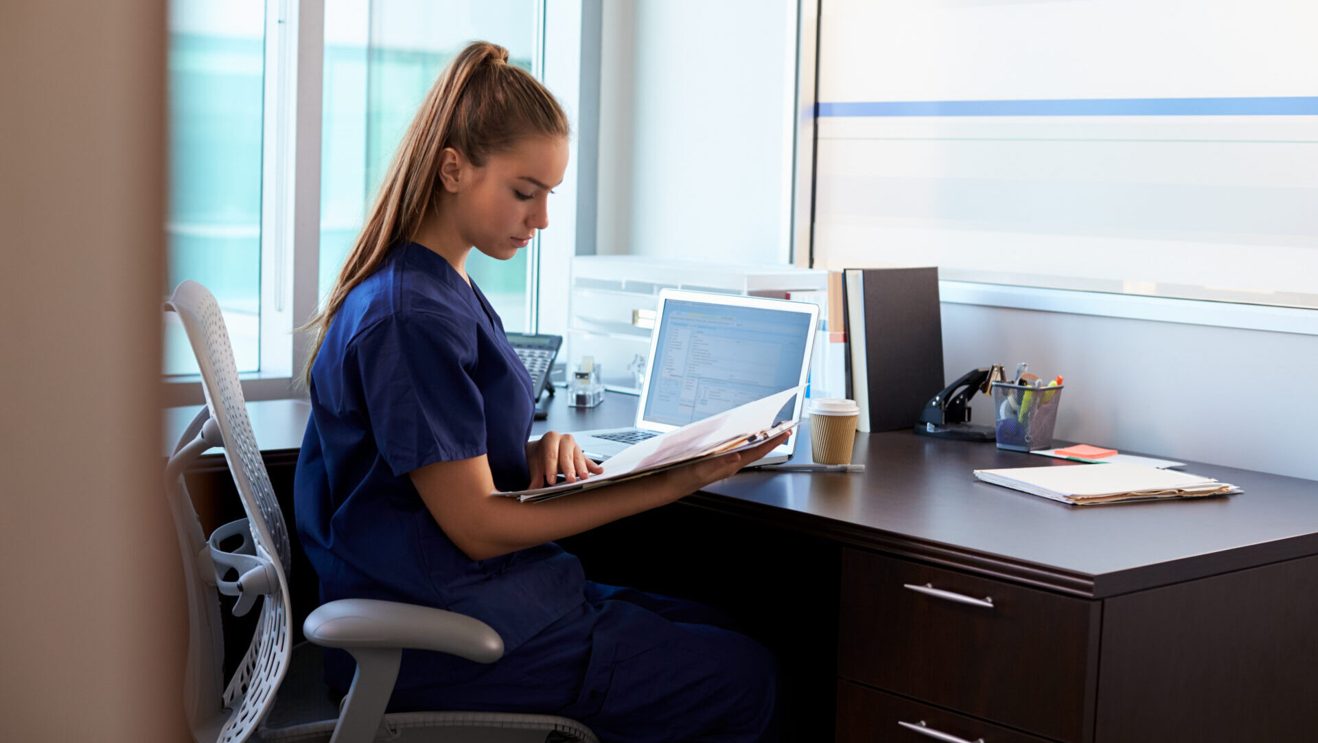 Woman in blue scrubs sitting at an office desk reviewing documents with a laptop and coffee cup nearby.