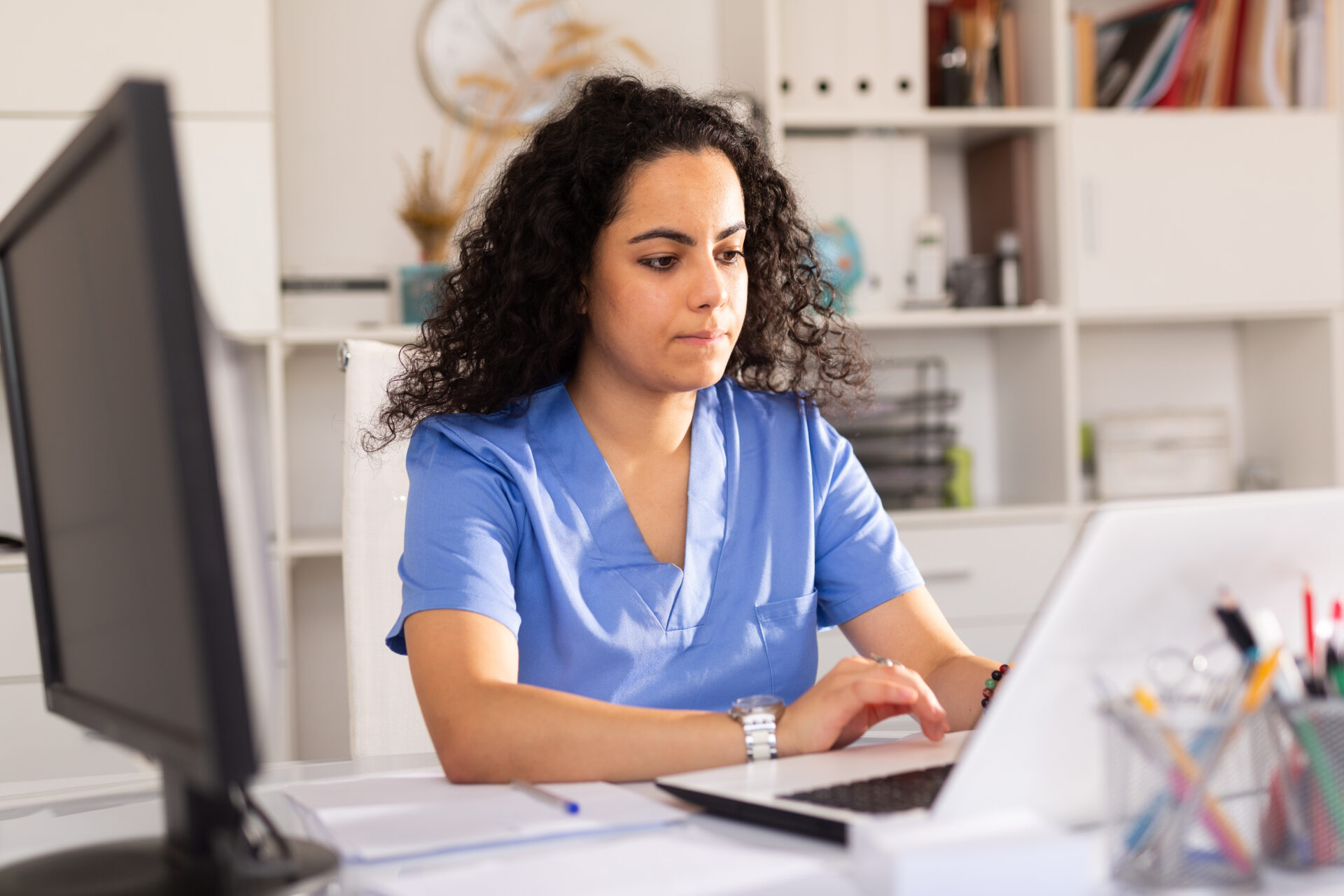 Woman in blue scrubs working on a laptop at a desk with office supplies nearby.