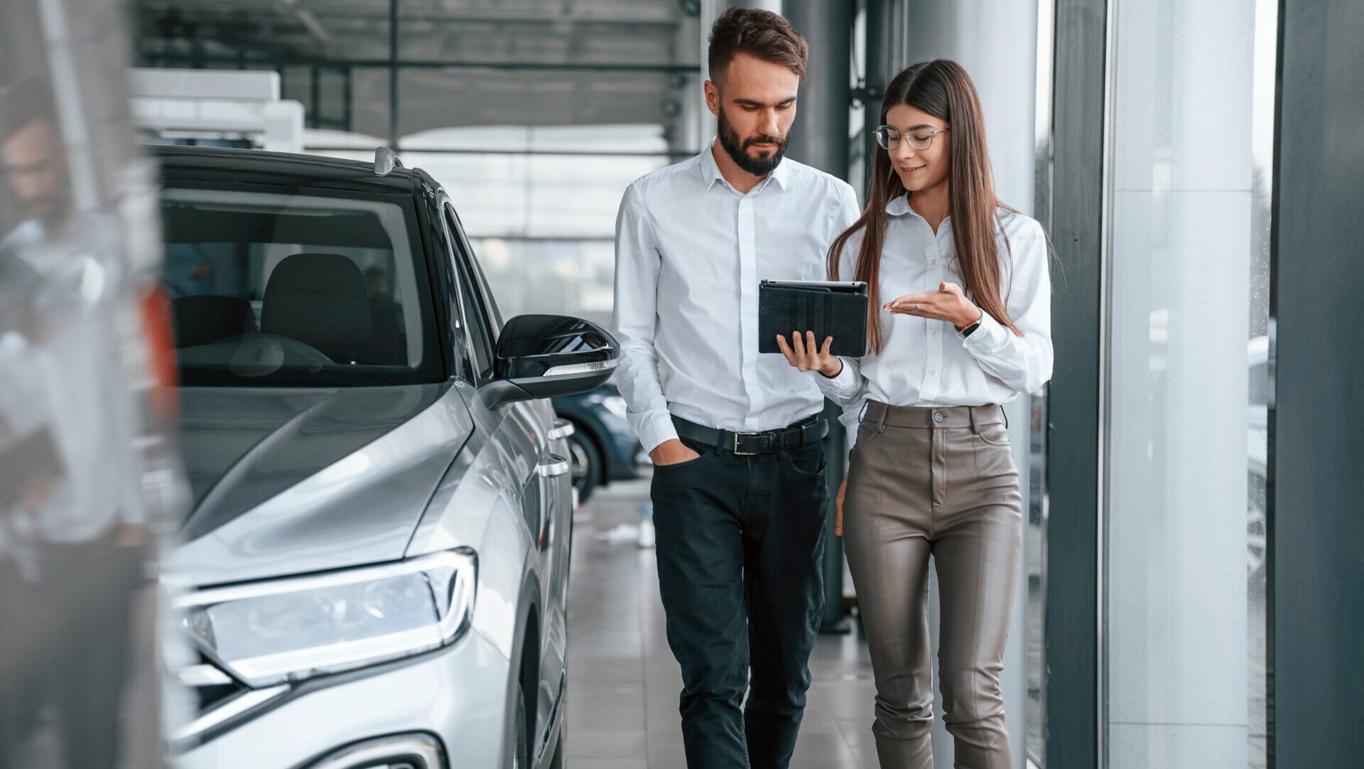 Man and woman in white shirts looking at a tablet while standing near a silver car indoors.