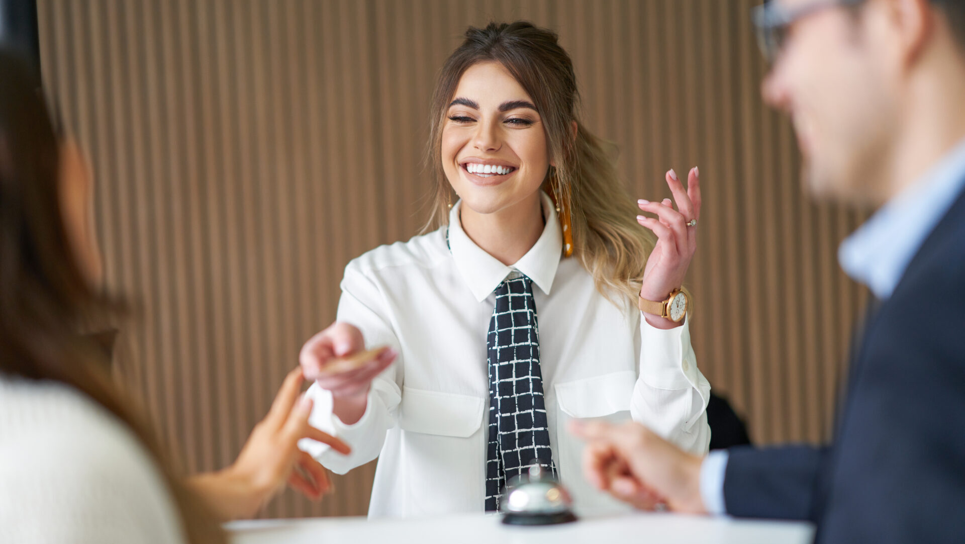 Smiling receptionist in white shirt handing a keycard to a guest at a hotel front desk.