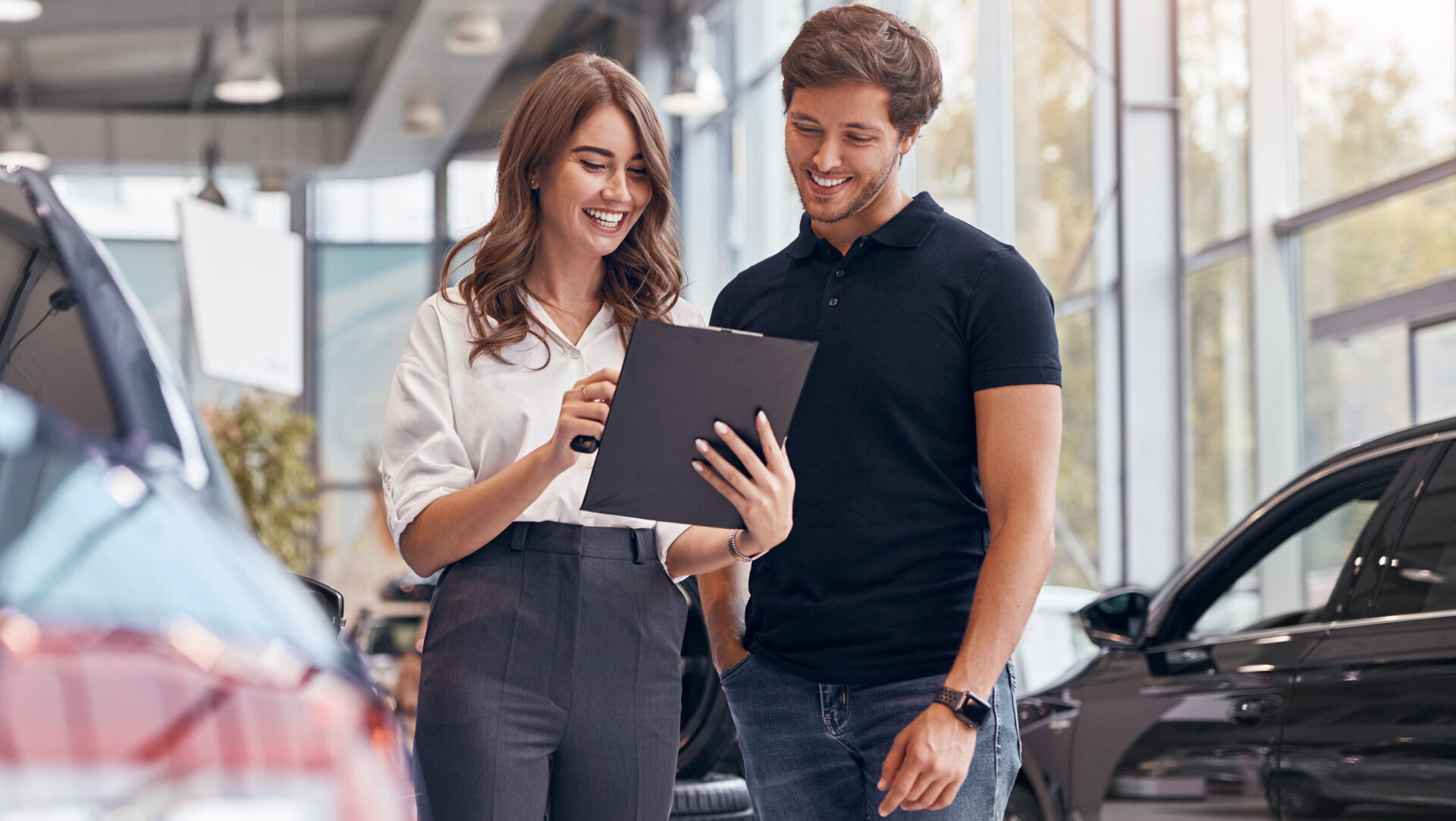 A woman showing a clipboard to a man in a car dealership with cars around them.