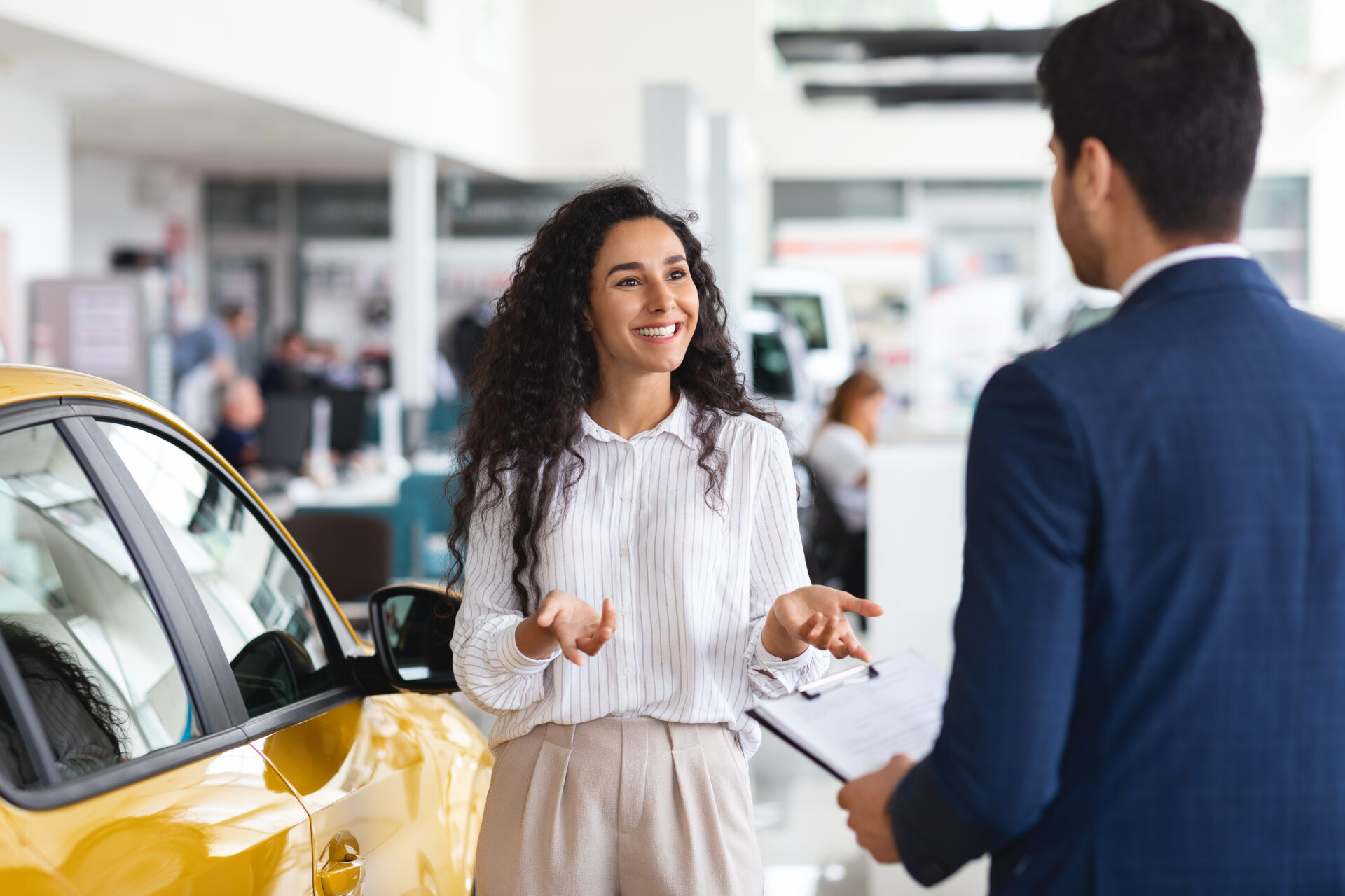 Smiling woman talking with a salesman near a yellow car in a dealership showroom.
