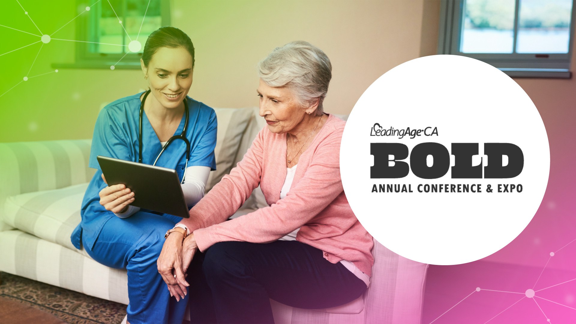 A nurse in blue scrubs shows a tablet to an elderly woman sitting on a couch at home.