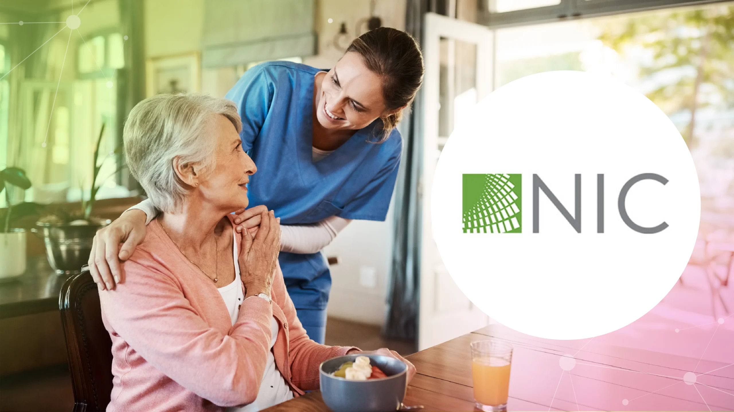 Smiling caregiver in blue scrubs embracing an elderly woman in pink at a dining table with food and juice.