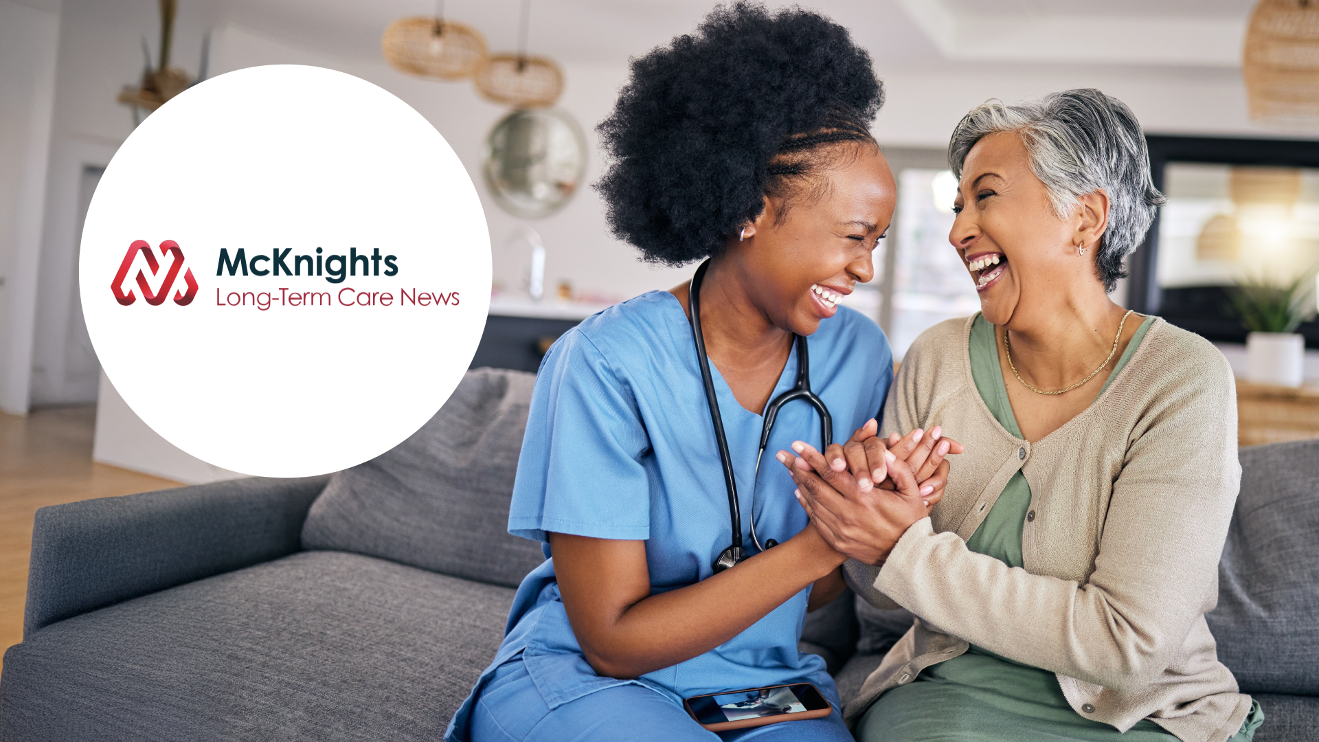 Smiling nurse in blue scrubs holding hands and laughing with elderly woman on a couch.
