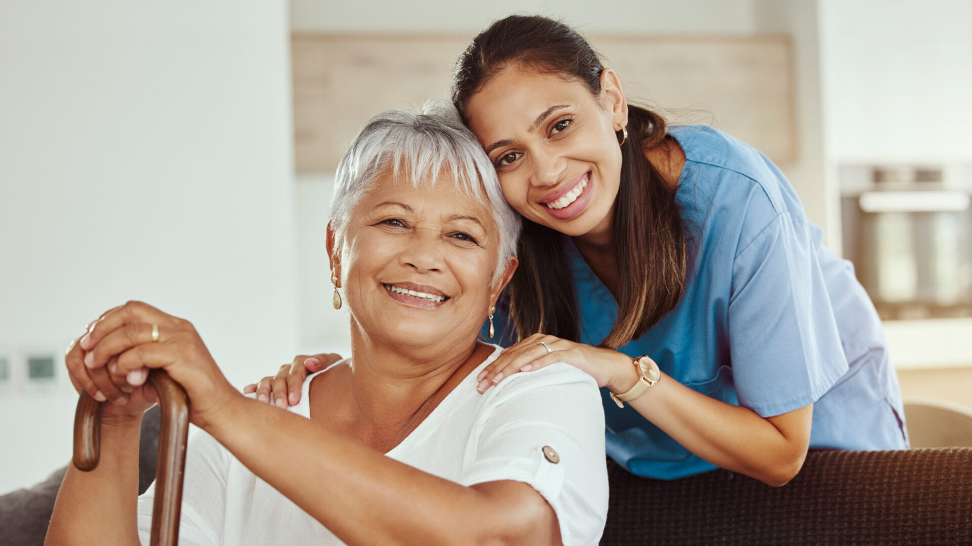 Smiling elderly woman with a cane and a caregiver in blue scrubs embracing her from behind.