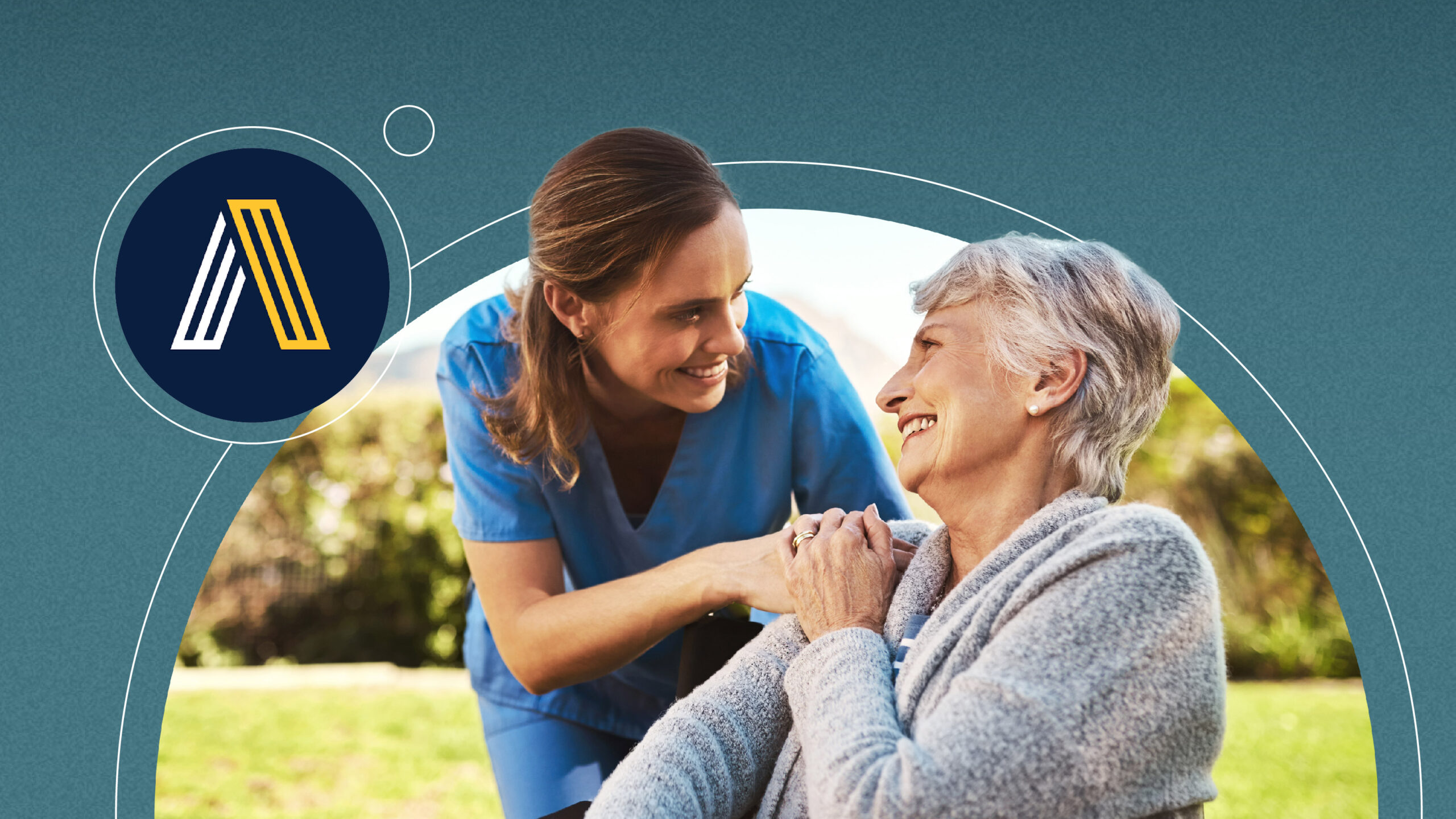 Caregiver in blue scrubs smiling and holding hands with elderly woman outdoors in a wheelchair.