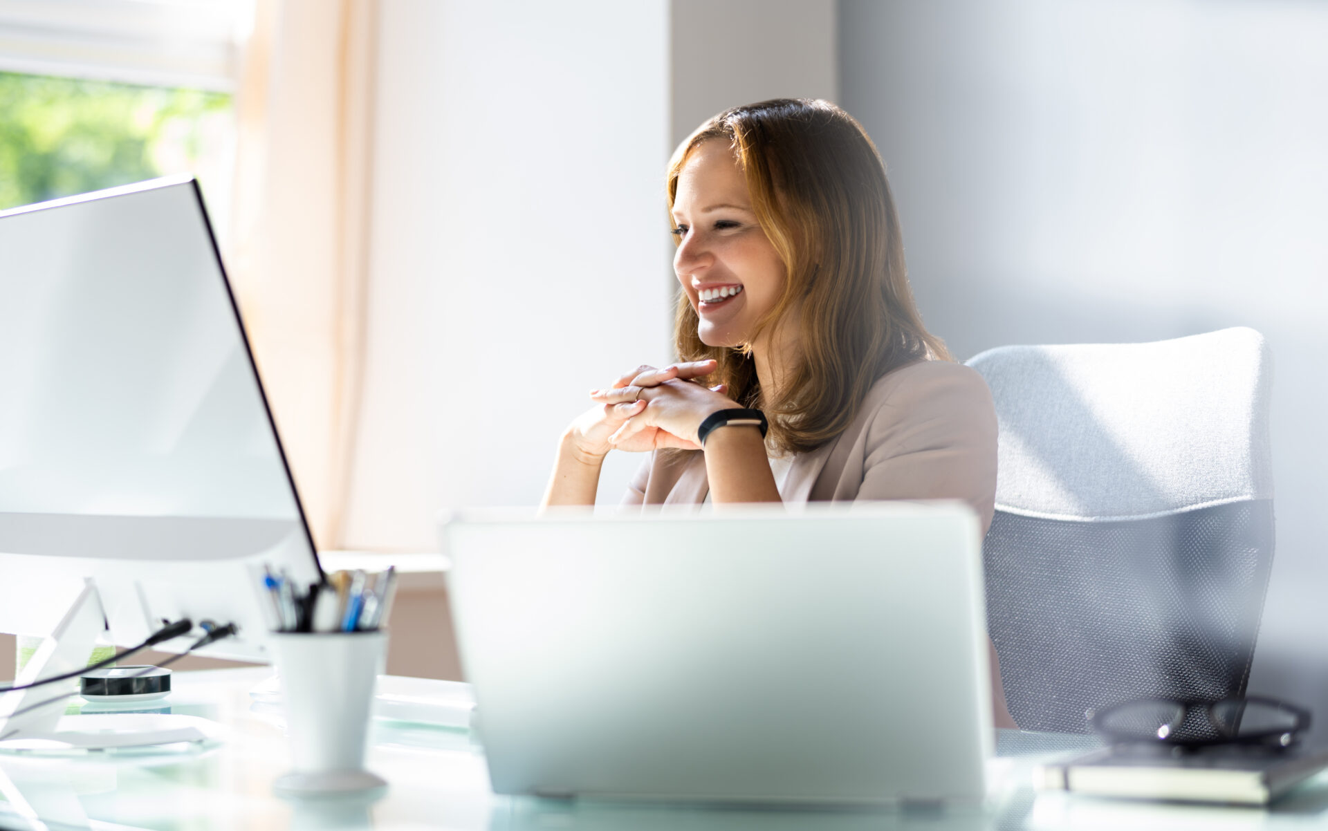 Smiling woman in office looking at a desktop monitor with a laptop and pen holder in foreground.