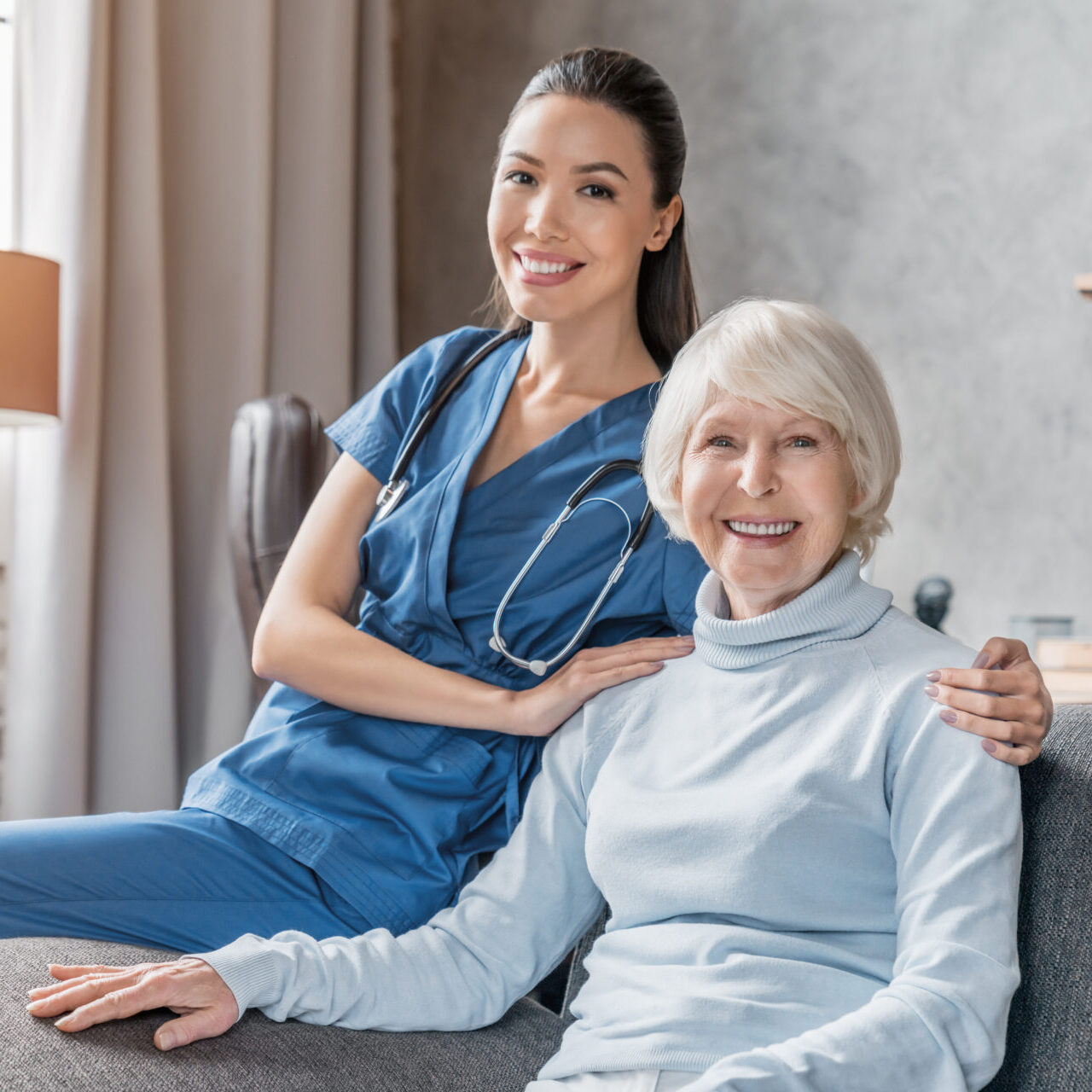 Smiling nurse in blue scrubs with stethoscope sitting beside happy elderly woman on couch.