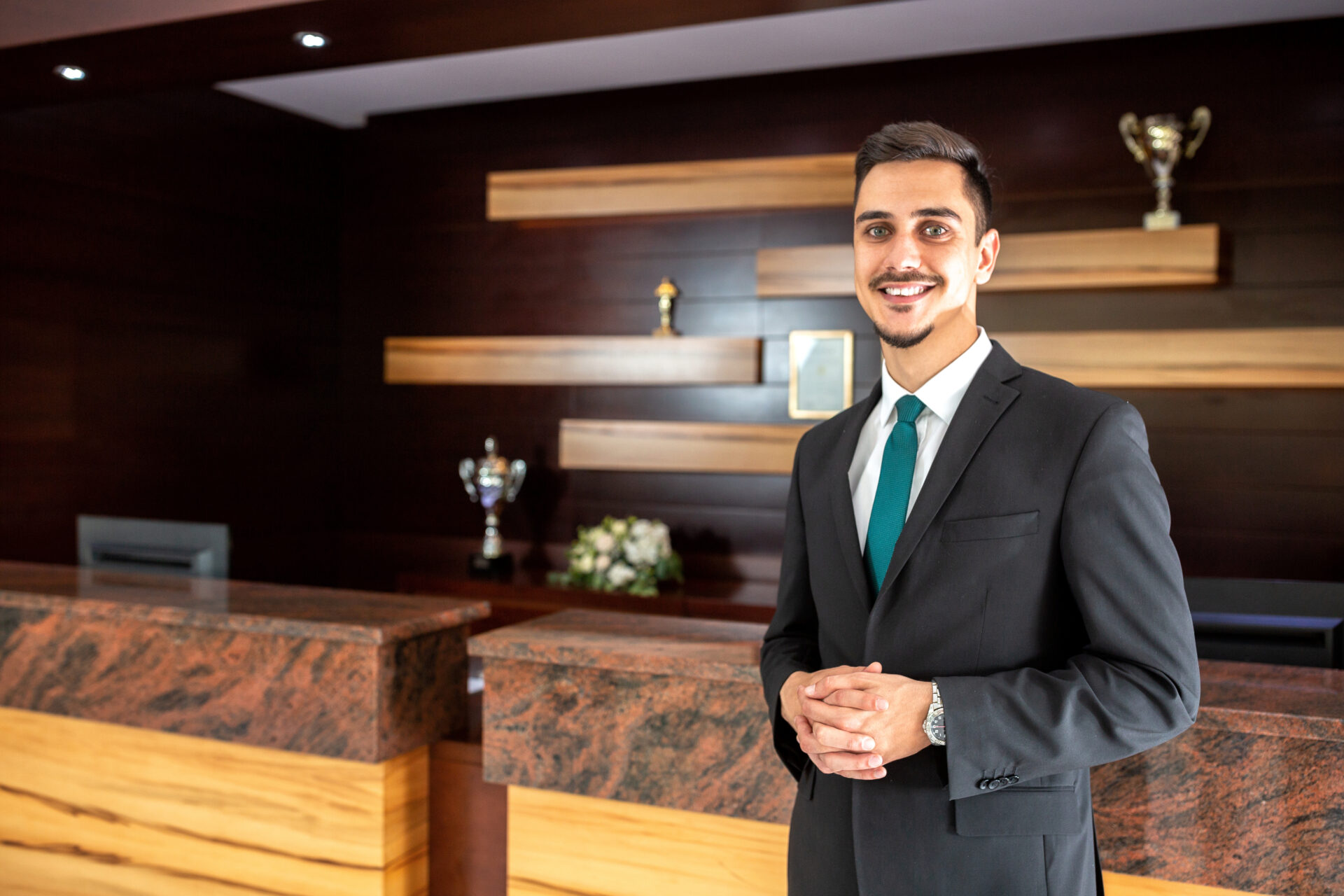 Smiling man in suit and teal tie standing in a modern hotel or office reception area.