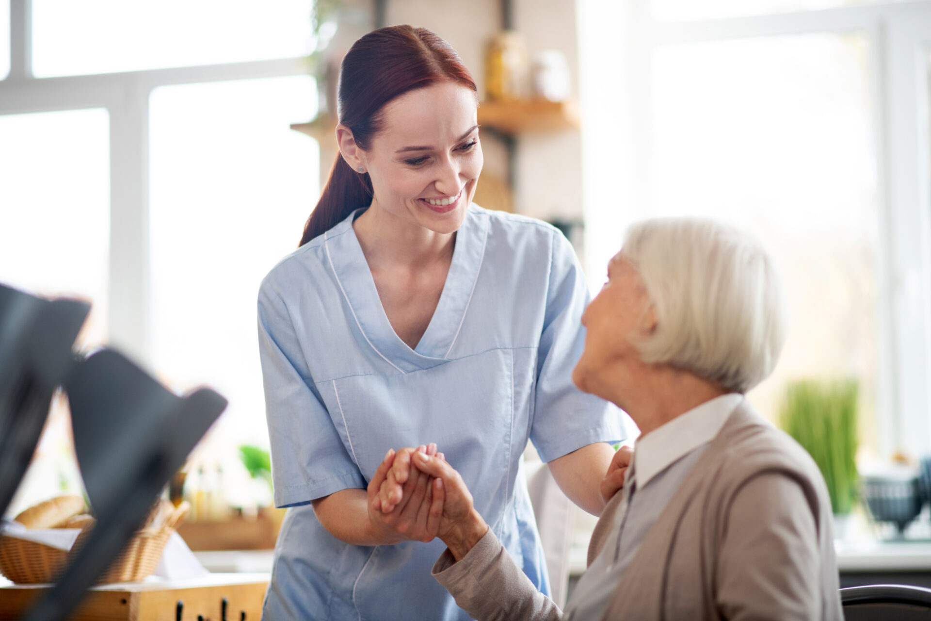 Caregiver in blue scrubs holding hands and smiling at an elderly woman in a wheelchair.
