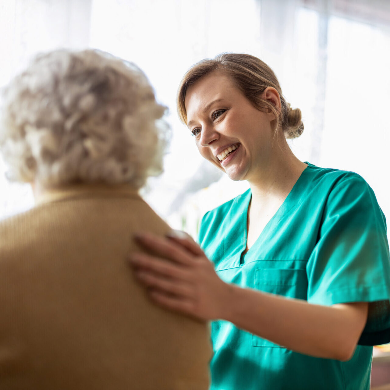 Smiling healthcare worker in scrubs comforts elderly person by gently placing a hand on their shoulder.