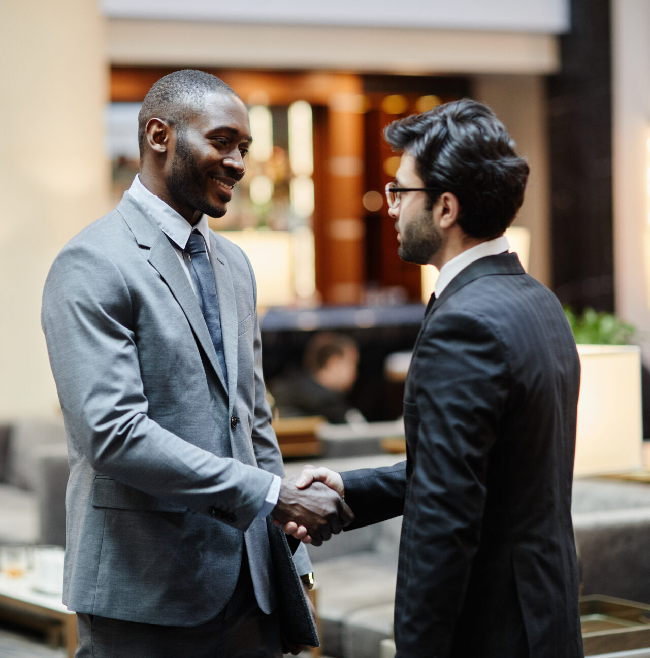 Two businessmen in suits shaking hands in a modern office lounge area.