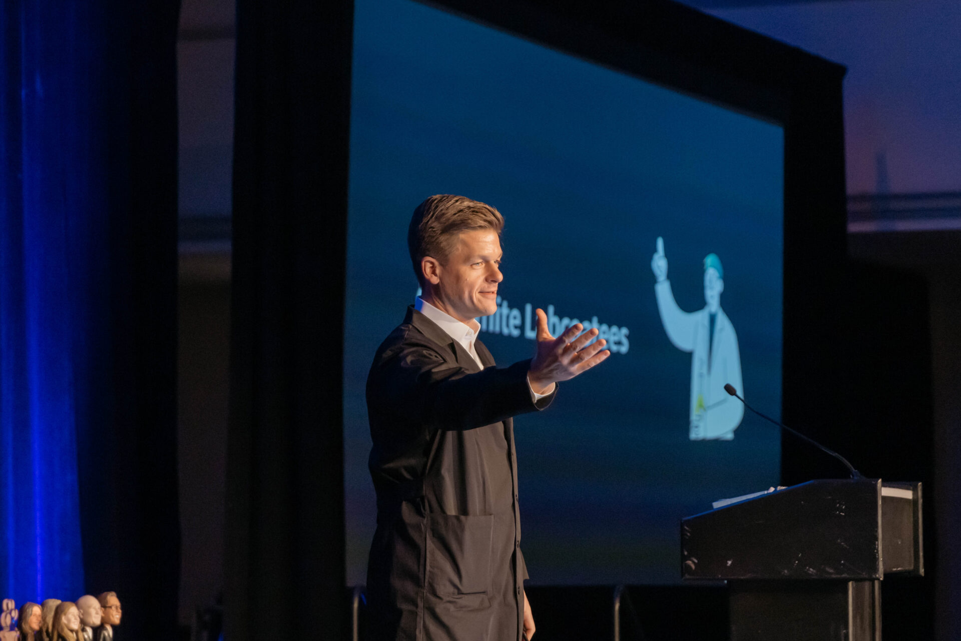 Man in dark coat speaking and gesturing with outstretched hand at a podium during a presentation.