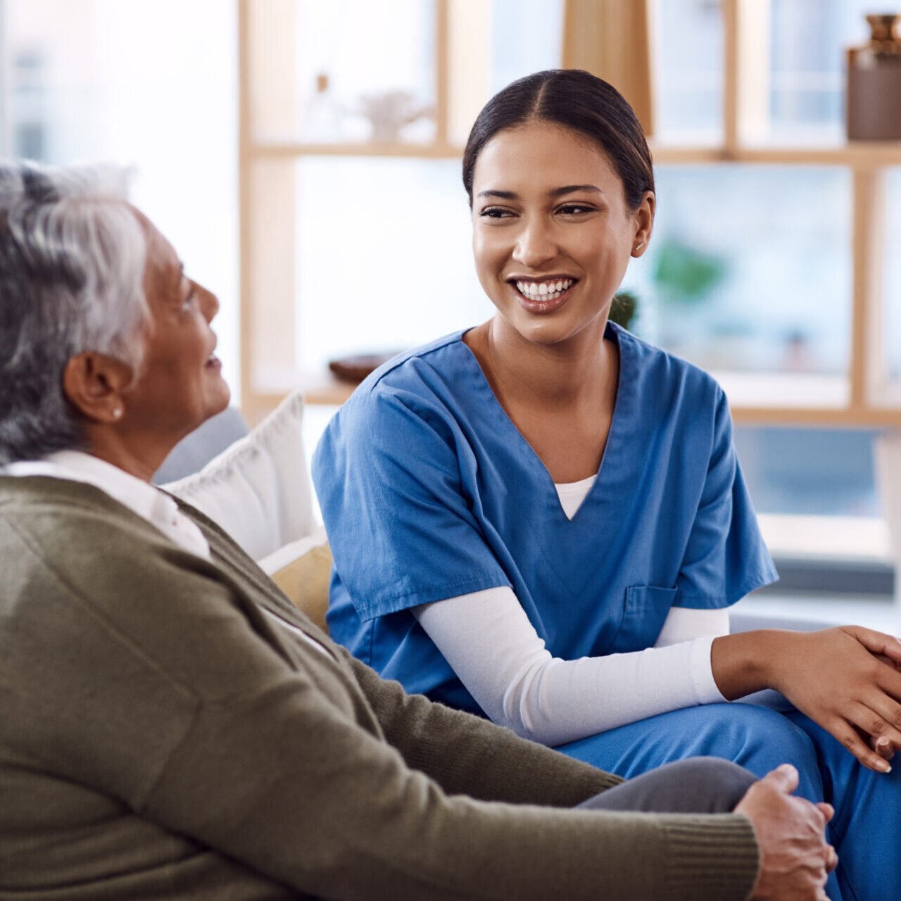 Smiling healthcare worker in blue scrubs talks with elderly woman sitting on a couch.