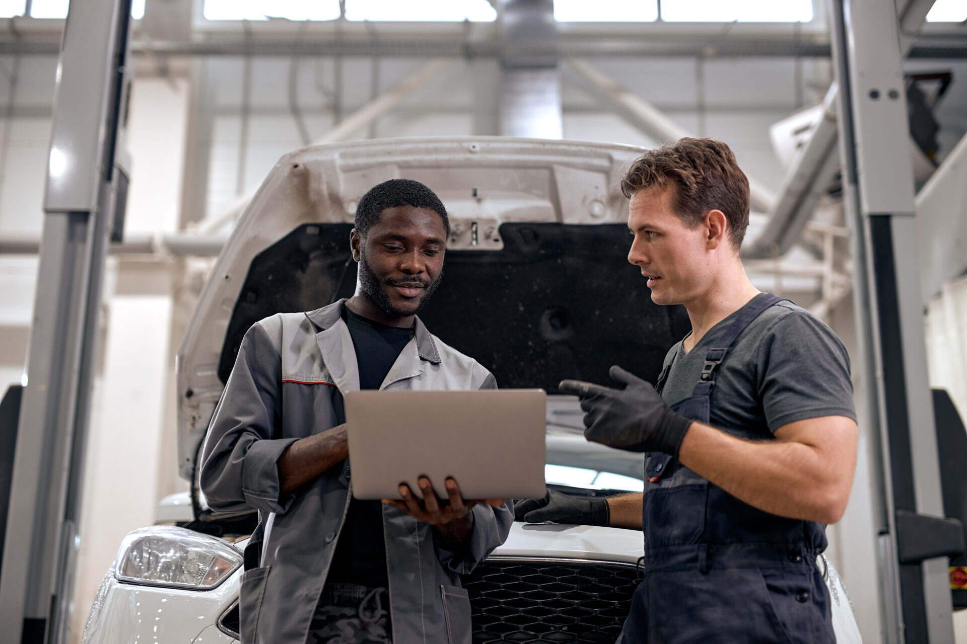 Two male mechanics stand by a car with its hood open, looking at a laptop in a garage.