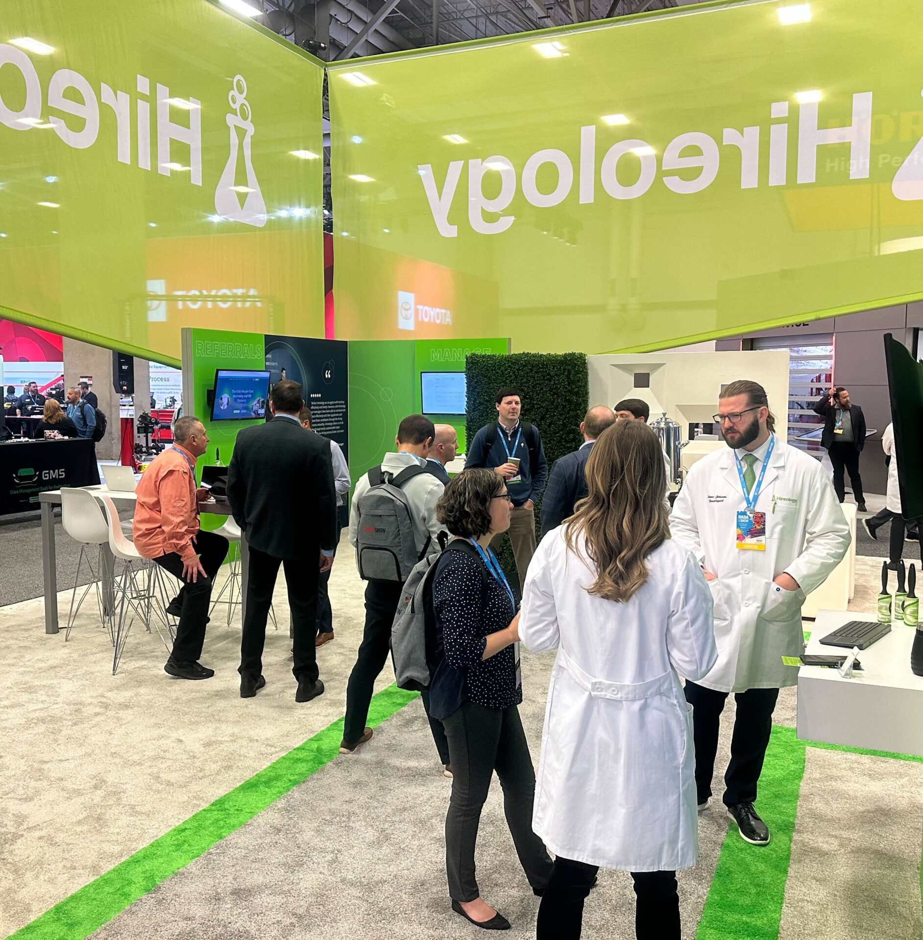 People talking and interacting inside a trade show booth with green accents and large hanging signs.