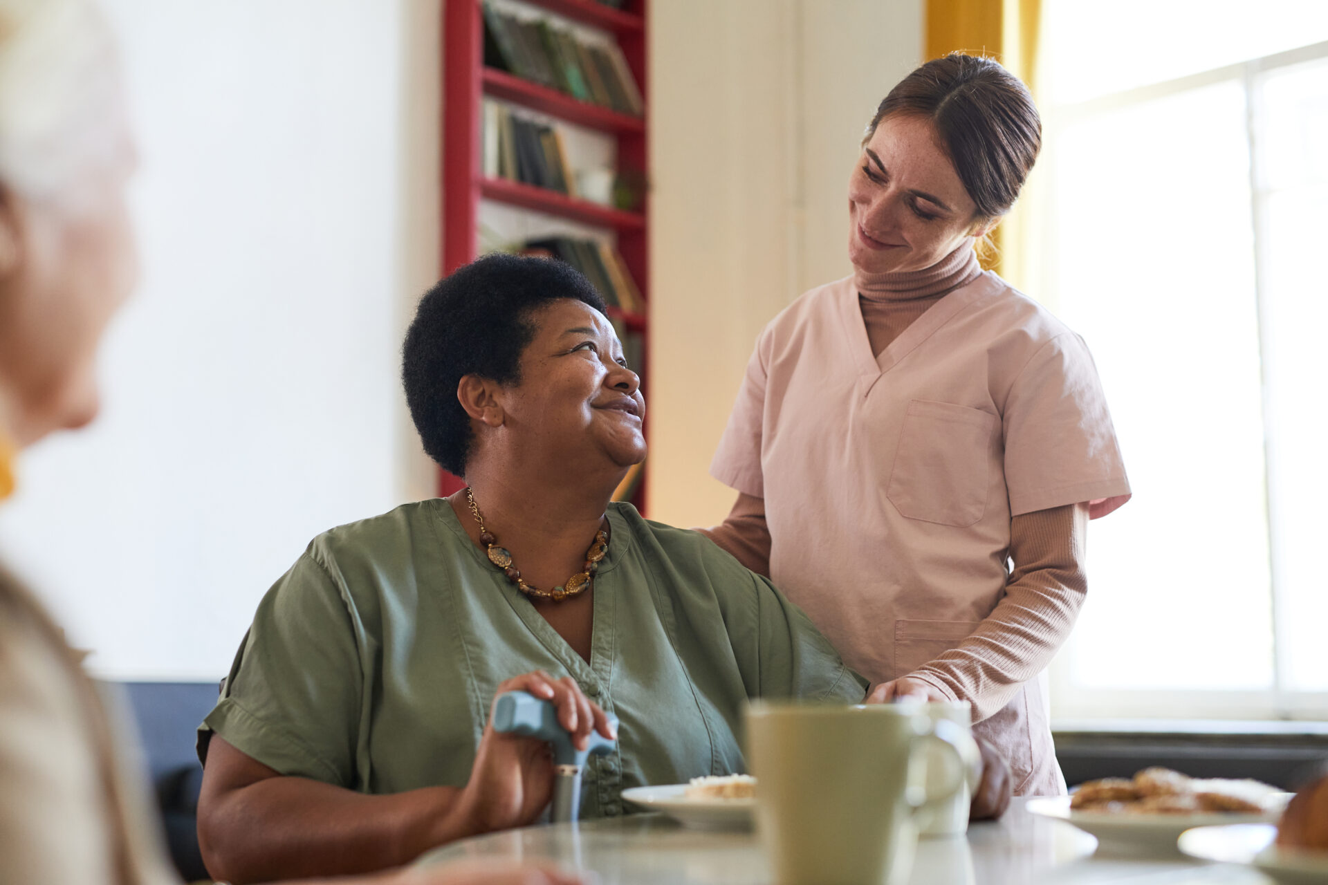 Caregiver in scrubs smiling and comforting a seated woman with a walking cane at a table.