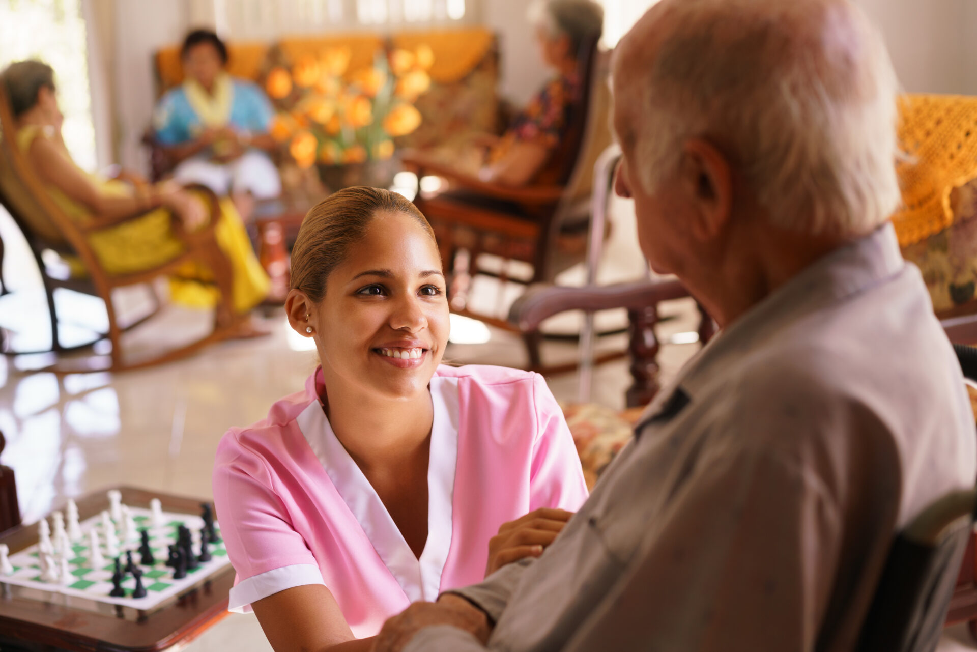 Young caregiver in pink uniform smiling warmly at an elderly man in a wheelchair inside a nursing home.