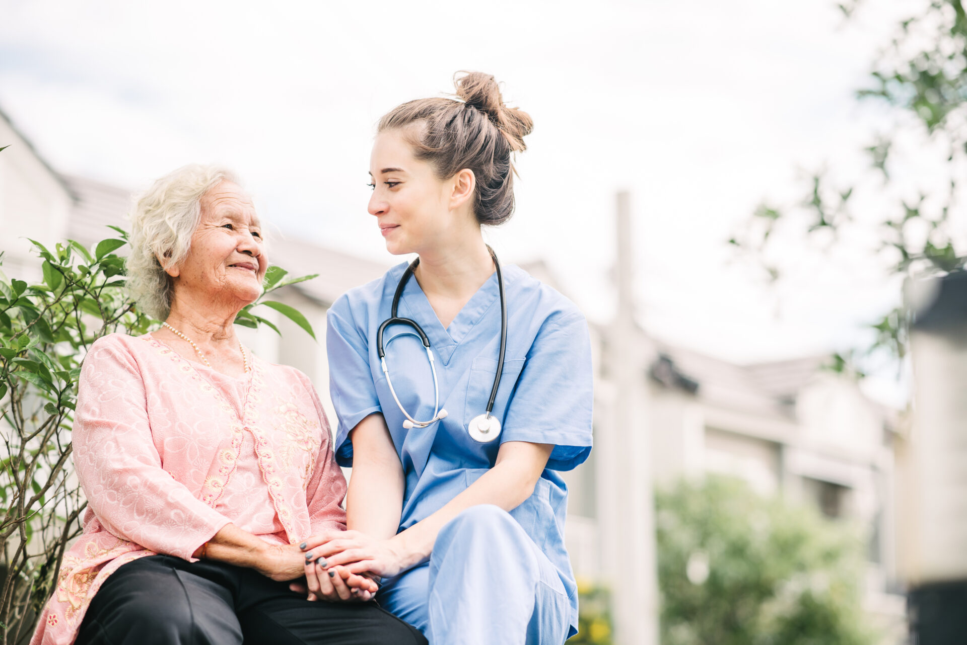 Young nurse in scrubs holding hands and smiling with elderly woman sitting outdoors.