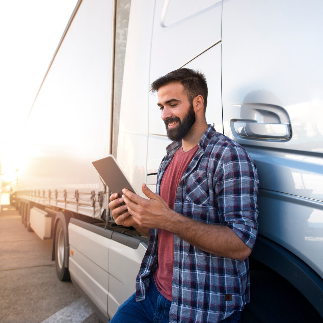 Bearded man in plaid shirt leaning on white truck while smiling and using a digital tablet outdoors.