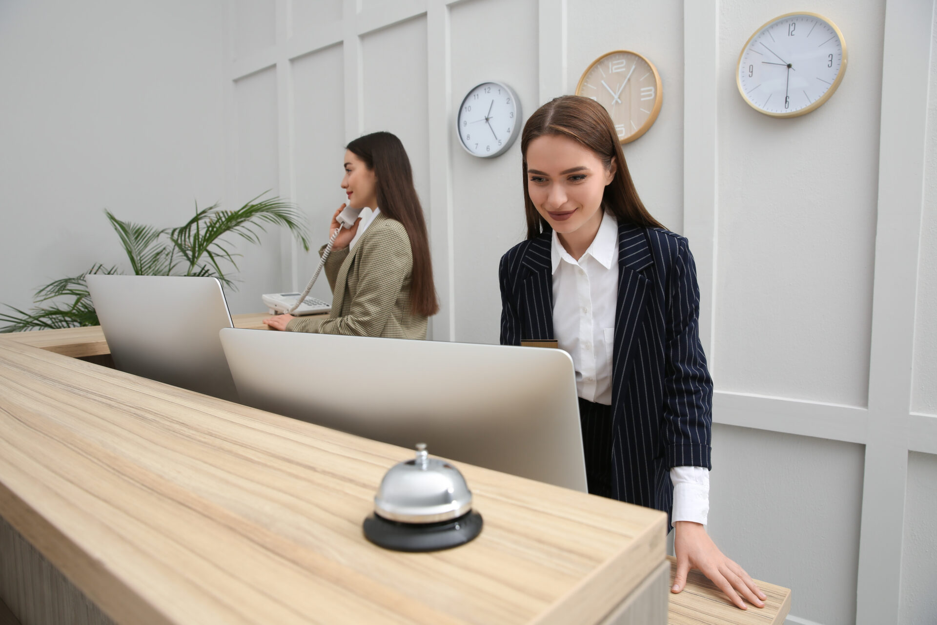 Two receptionists working behind a wooden front desk with clocks on the wall and a service bell.