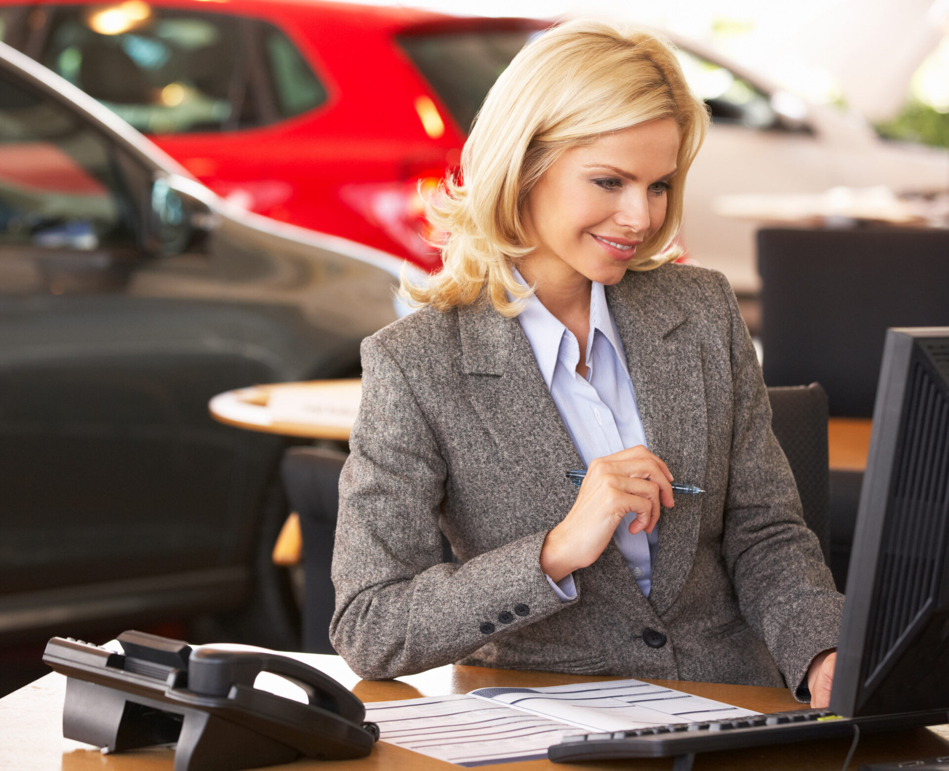 Woman in gray blazer working at a desk with computer and phone, cars in background.