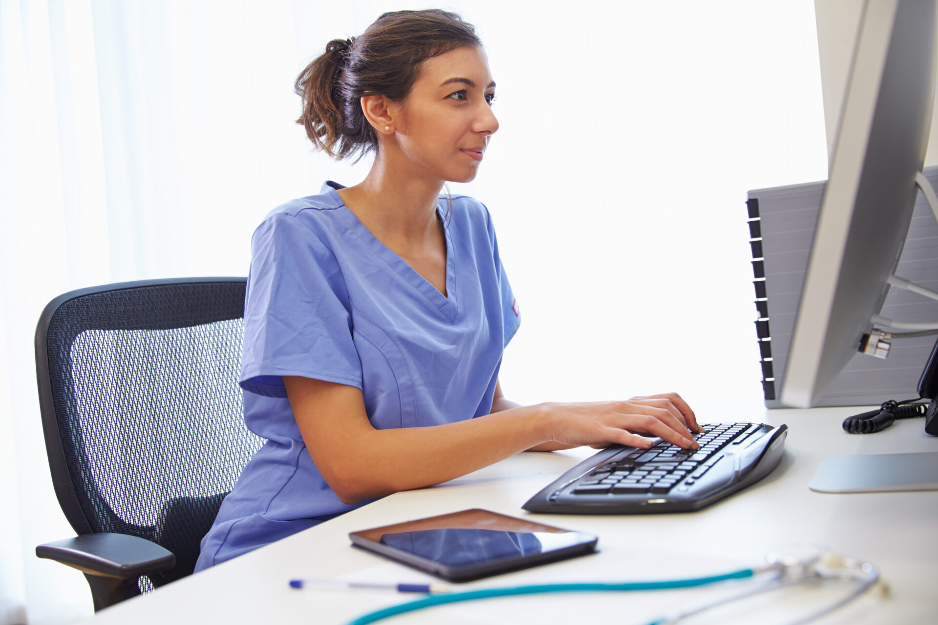 Healthcare worker in blue scrubs typing on a keyboard at a desk with a tablet and stethoscope nearby.