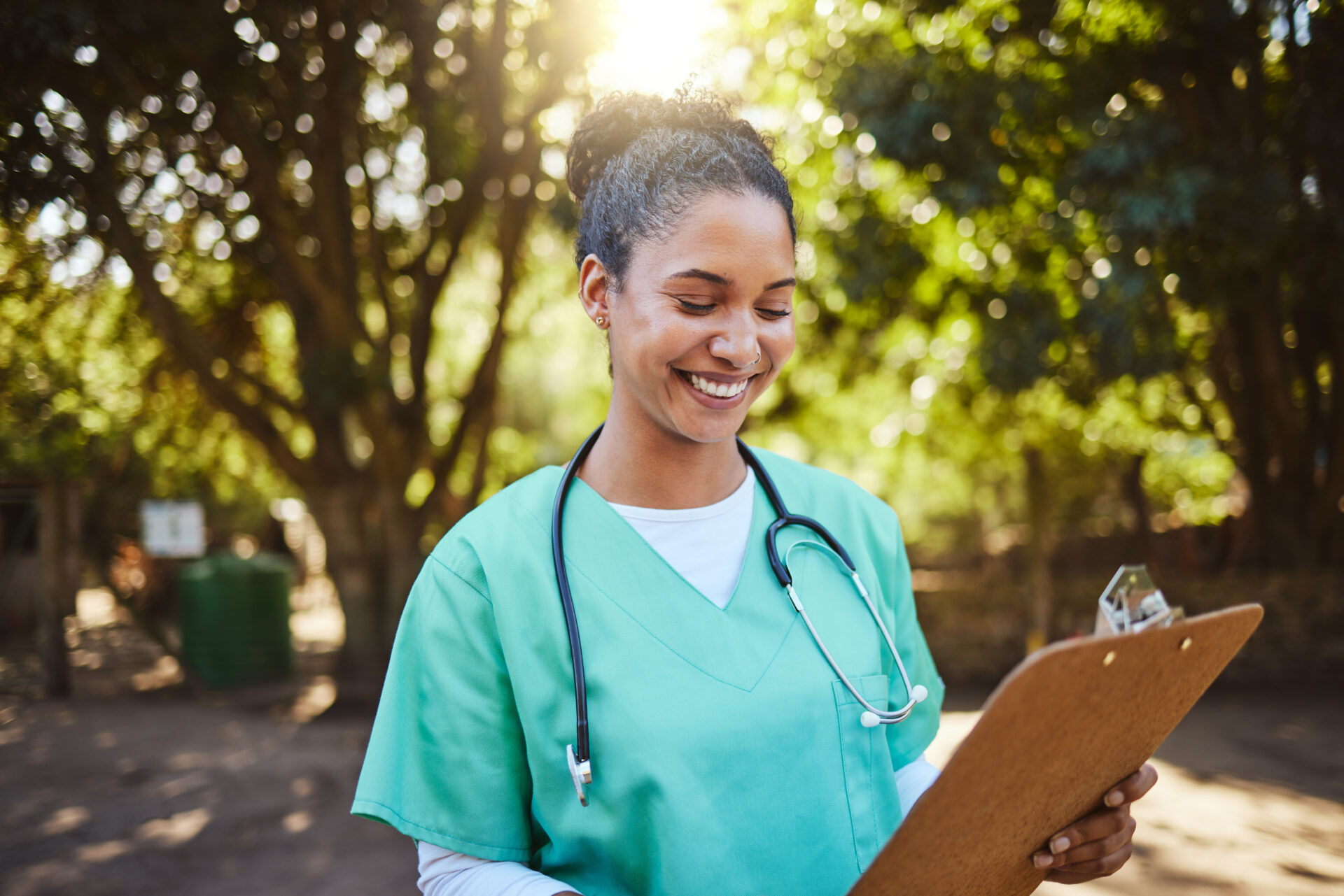 Smiling female healthcare worker in green scrubs with stethoscope holding a clipboard outdoors.