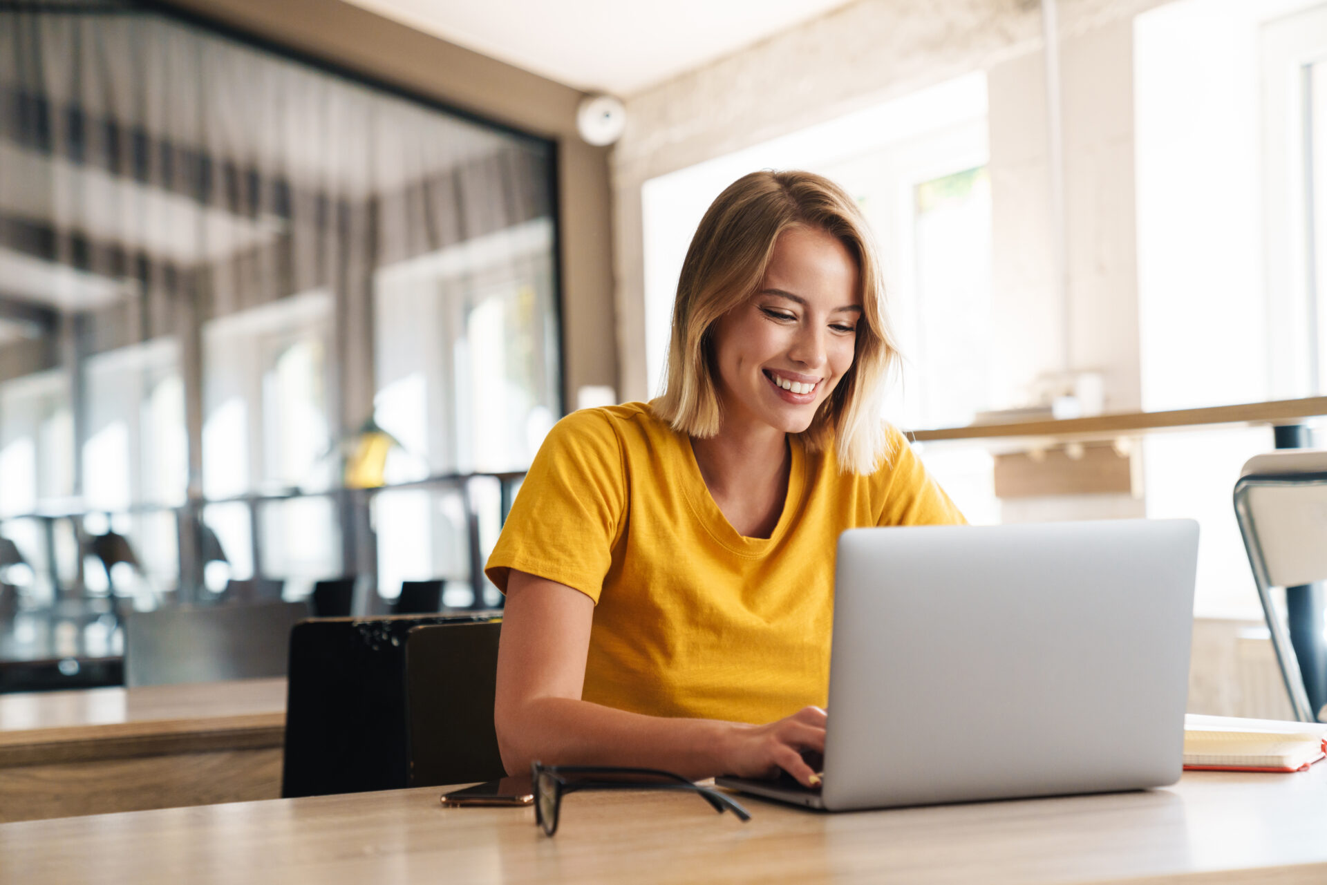 Smiling woman in yellow shirt working on a laptop at a bright modern office desk.