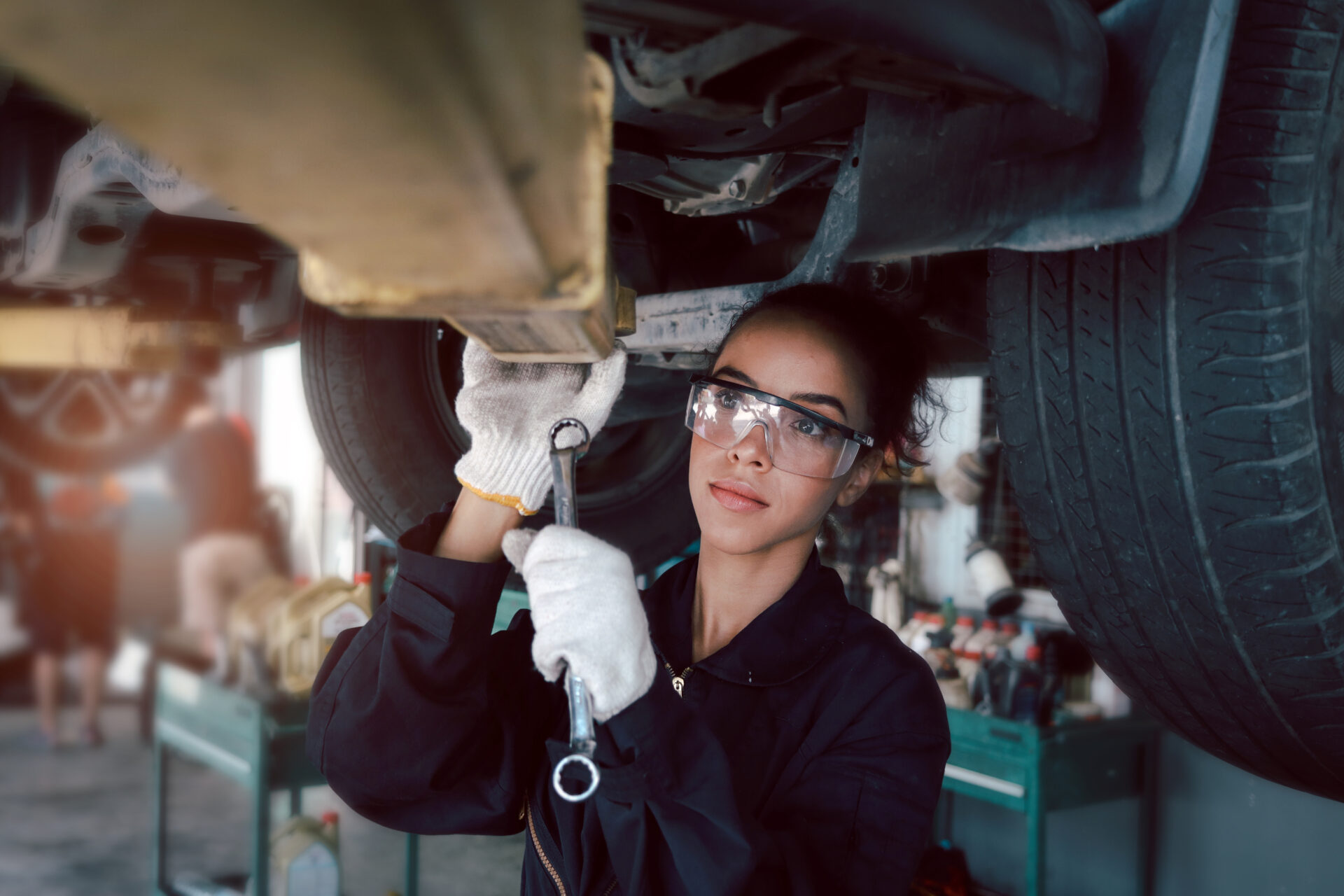 Young woman mechanic with safety glasses and gloves working under a raised vehicle with a wrench.