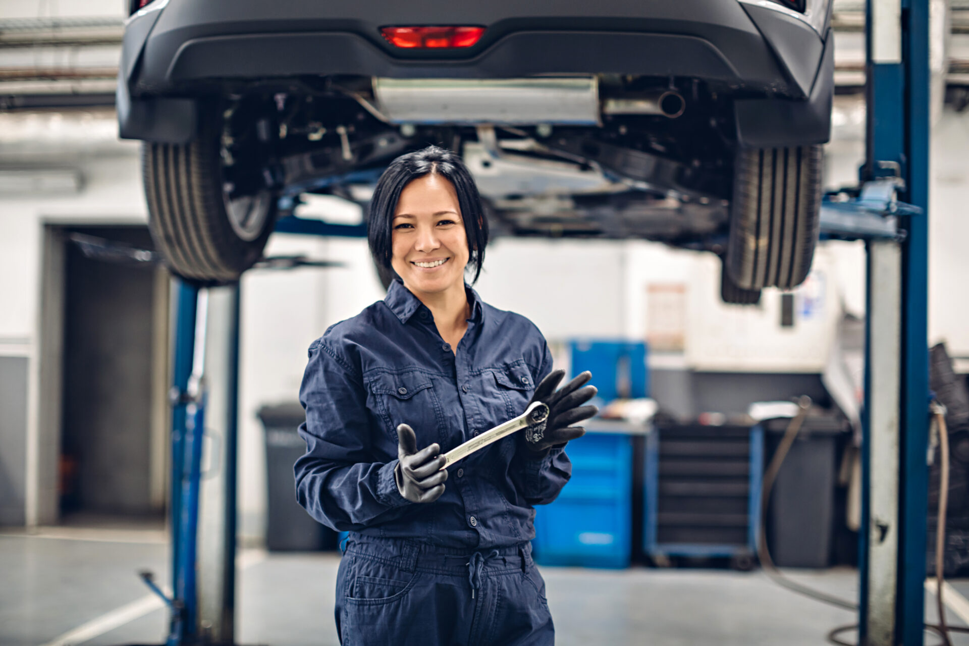 Smiling female mechanic holding a wrench under a car lifted on a hydraulic lift in a garage.