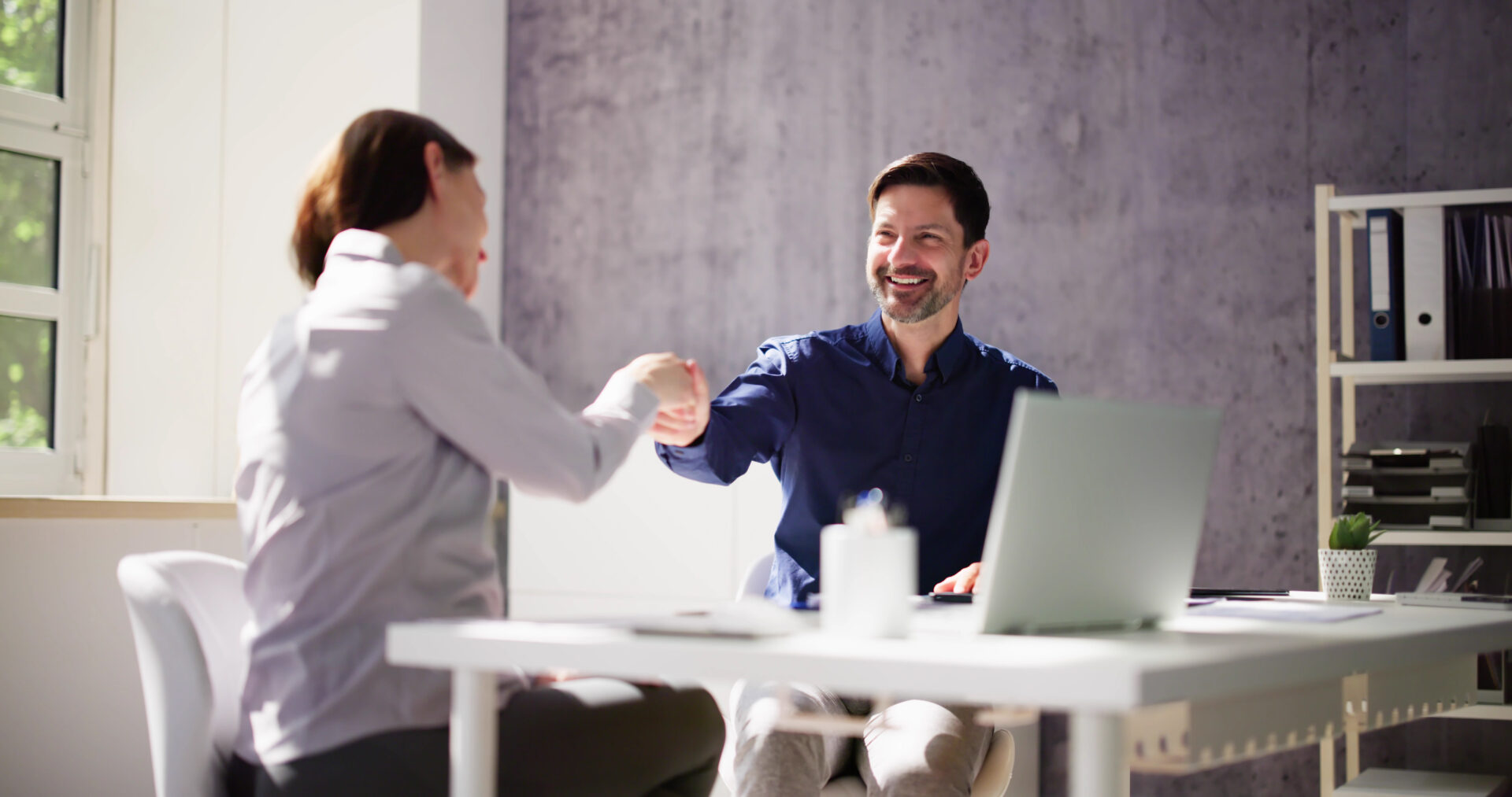 Two people shaking hands across a white office desk with a laptop and shelves in the background.
