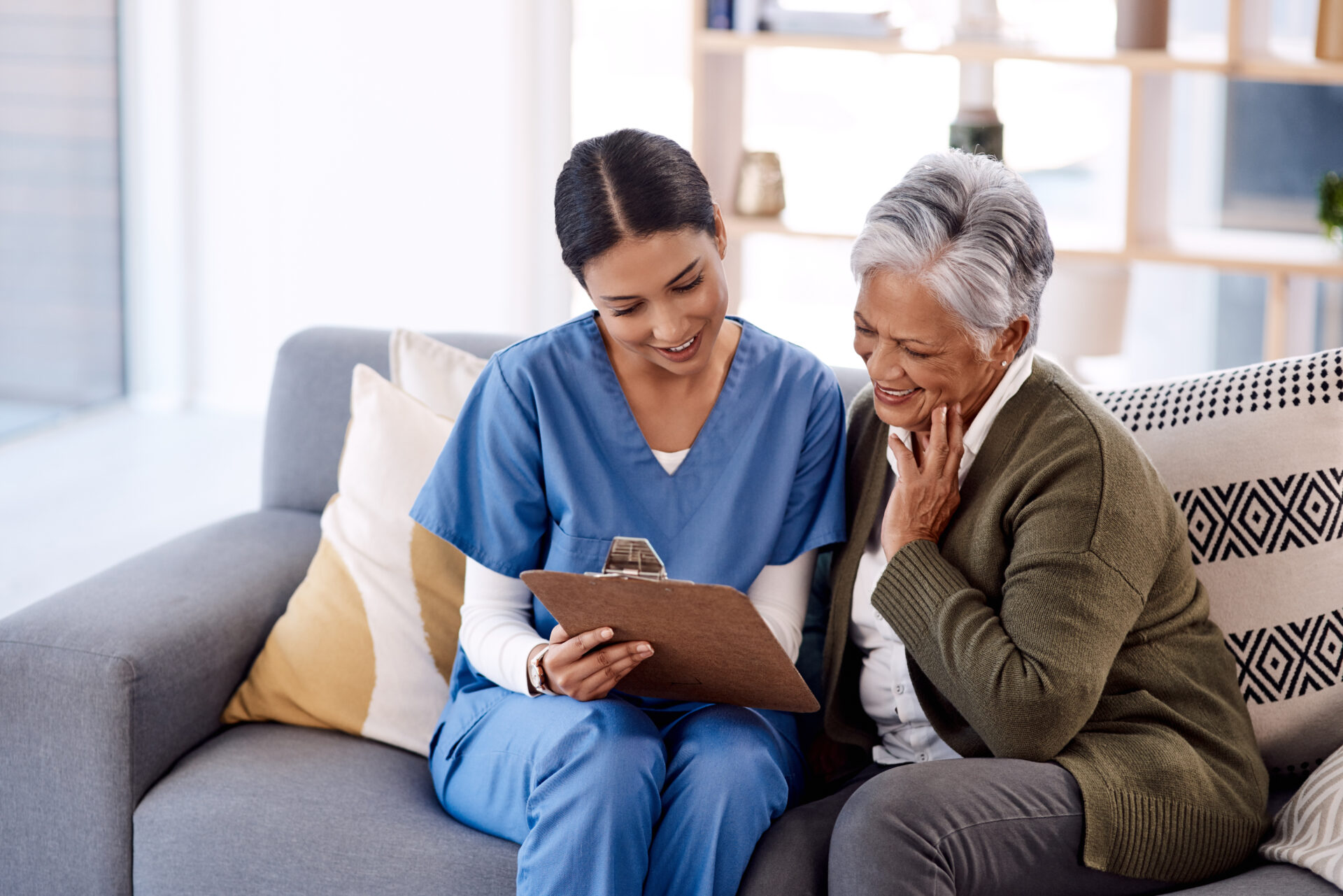Young nurse in blue scrubs shows clipboard to smiling elderly woman on a sofa in a cozy room.
