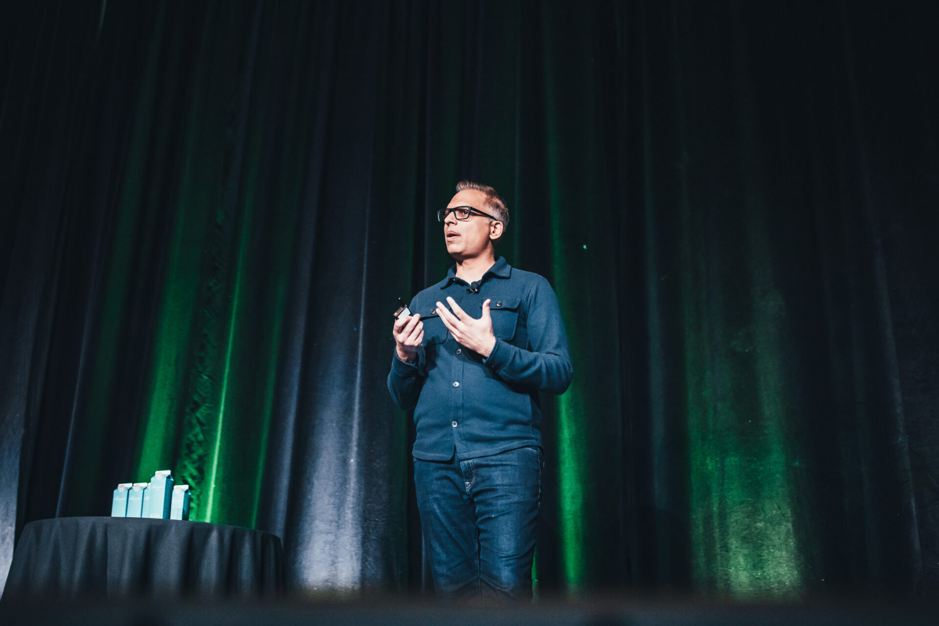 Man wearing glasses and blue jacket speaking on stage with green-lit black curtains behind him.