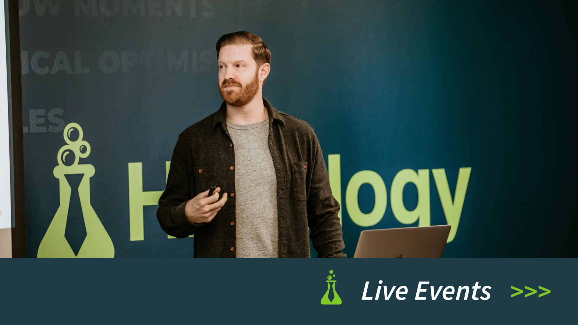 Man with red beard holding a clicker giving a presentation in front of a blue-green background with text and a laptop.